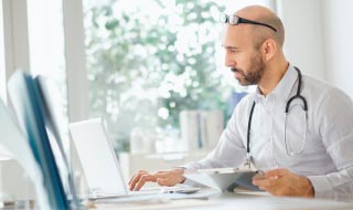 Médico (hombre) en su mesa de la consulta mirando a una pantalla de ordenador